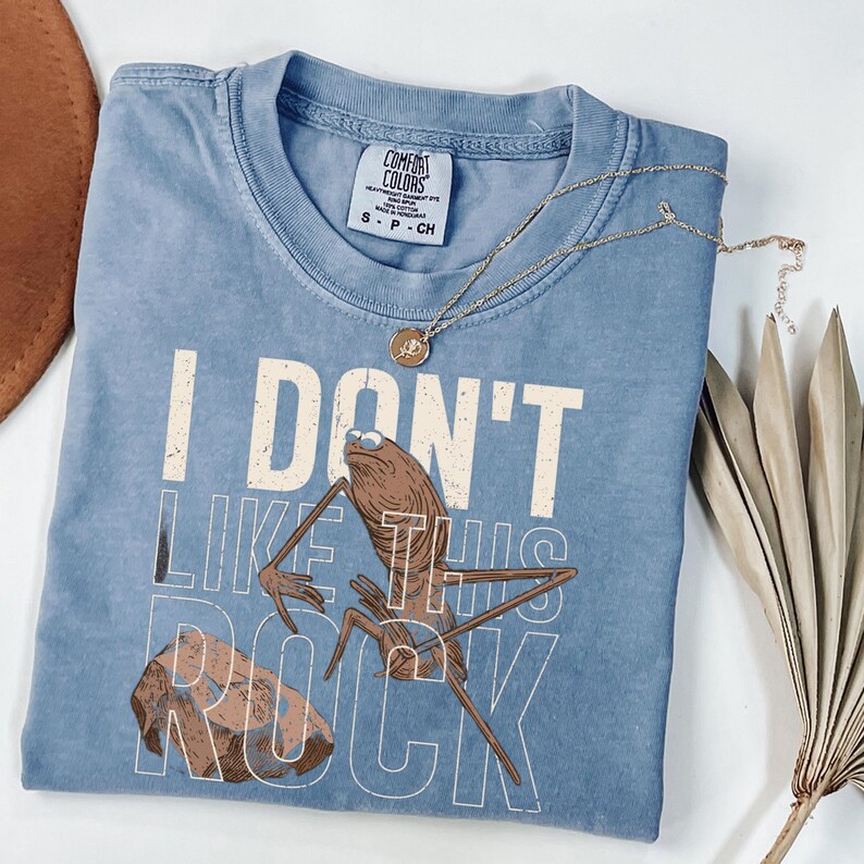 May include: A light blue Comfort Colors t-shirt with the text "I DON'T LIKE THIS ROCK" and a cartoon illustration of a creature with sunglasses. A gold necklace with a pendant is draped across the shirt.