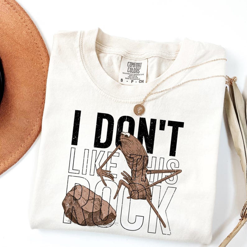 May include: Cream-colored t-shirt with the text "I DON'T LIKE HIS ROCK" in black and white. The graphic features a cartoonish insect with a rock. A gold necklace and a brown hat are also visible.
