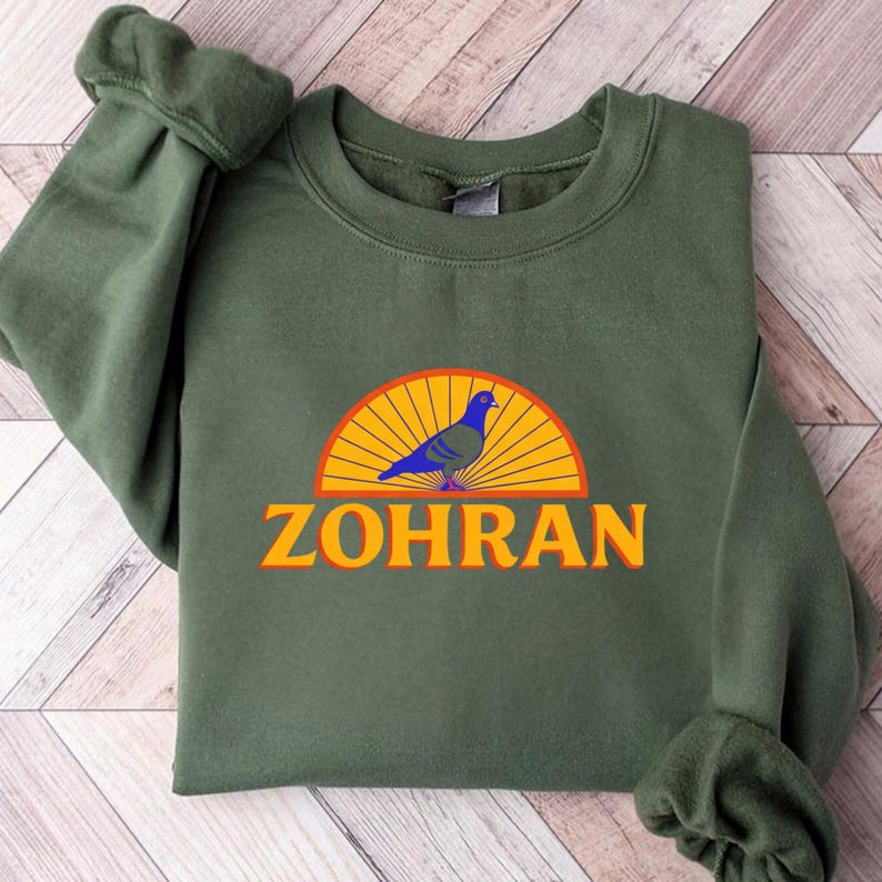 May include: An olive green sweatshirt featuring a graphic of a blue pigeon perched on a yellow and orange sunburst design. The name "ZOHRAN" is printed in orange below the graphic. The sweatshirt is made of a soft material.