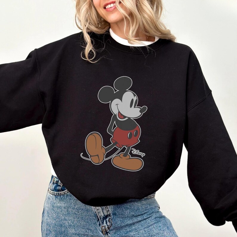 Puede incluir: Sudadera negra con un gr&aacute;fico de Mickey Mouse. El personaje de dibujos animados se muestra en su pose cl&aacute;sica, con pantalones cortos rojos, zapatos marrones y guantes blancos. El logotipo de Disney es visible en la parte inferior derecha.