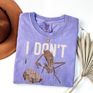 May include: A lavender Comfort Colors t-shirt with the phrase "I DON'T LIKE THIS ROCK" and a cartoon illustration of a bug-like creature. A gold necklace and a brown hat are also visible.