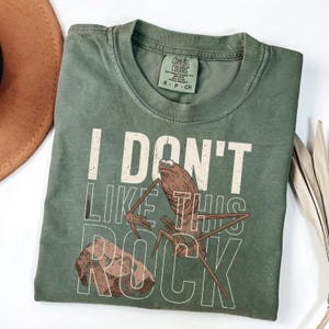 May include: Sage green t-shirt with the text "I DON'T LIKE THIS ROCK" and a cartoon illustration of a brown insect-like creature and a rock. The shirt has a Comfort Colors tag.
