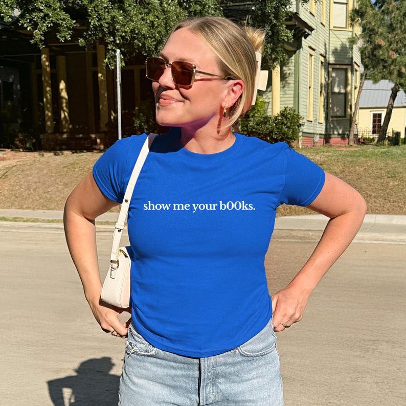 May include: A royal blue t-shirt with the text "show me your books" in white. The shirt is short-sleeved and appears to be made of a soft material. The person is wearing sunglasses and a white purse.