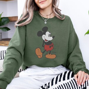 Puede incluir: Una sudadera verde oliva con un gr&aacute;fico de Mickey Mouse. El personaje de dibujos animados se muestra en su pose cl&aacute;sica, con pantalones cortos rojos, zapatos marrones y guantes blancos. El logotipo de Disney es visible debajo.