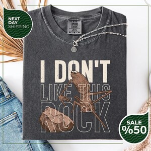 May include: Dark gray t-shirt with the text "I DON'T LIKE THIS ROCK" and a cartoon illustration of a bug and a rock. A silver necklace is draped across the shirt.