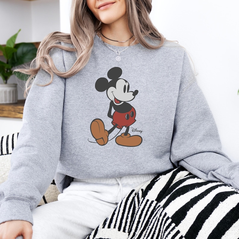 Puede incluir: Sudadera gris claro con un gr&aacute;fico de Mickey Mouse. El personaje de dibujos animados est&aacute; en negro, rojo y marr&oacute;n, con el logotipo de Disney. La sudadera es de estilo casual.