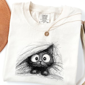 May include: Cream-colored t-shirt featuring a black, scribbled illustration of a cat with large eyes. The shirt has a crew neck and short sleeves. A gold necklace with a pendant is draped across the shirt. The tag reads "COMFORT COLORS".