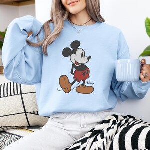Puede incluir: Sudadera azul claro con un gr&aacute;fico de Mickey Mouse. El personaje de dibujos animados se representa con su atuendo cl&aacute;sico, con orejas negras, pantalones cortos rojos y zapatos marrones. El logotipo de Disney es visible. La sudadera es de color azul claro liso.