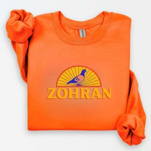 May include: Bright orange sweatshirt with a graphic featuring a blue and tan pigeon inside a yellow and orange semi-circle, with the name "ZOHRAN" in yellow block letters below. The sleeves are rolled up.