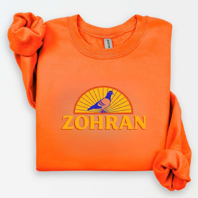 May include: Bright orange sweatshirt with a graphic featuring a blue and tan pigeon inside a yellow and orange semi-circle, with the name "ZOHRAN" in yellow block letters below. The sleeves are rolled up.