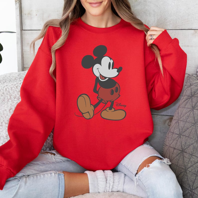 Puede incluir: Sudadera roja con un dise&ntilde;o de Mickey Mouse. El personaje de dibujos animados se muestra en su pose cl&aacute;sica, con pantalones cortos rojos y zapatos marrones grandes. El logotipo de Disney es visible en la parte inferior derecha.