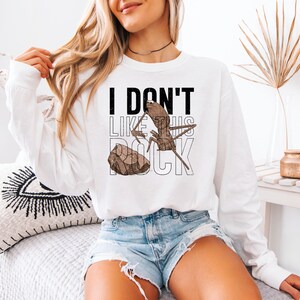 May include: White long-sleeve sweatshirt with the text "I DON'T LIKE THIS ROCK" in black and white. The graphic features a cartoon ant and a rock in brown tones. The sweatshirt is a casual, everyday wear item.