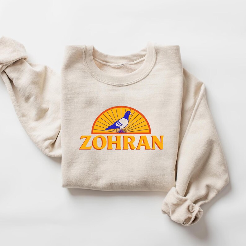 May include: Cream-colored sweatshirt with a crew neck and long sleeves. The front features a graphic of a blue pigeon inside a yellow and orange sunburst design, with the word "ZOHRAN" in orange block letters below.