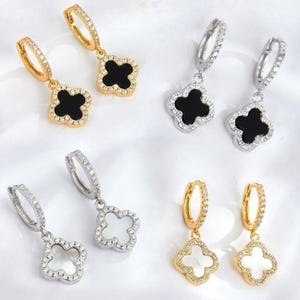 May include: Four pairs of hoop earrings featuring clover-shaped charms. Two pairs are gold-toned with black and white inlays, and two pairs are silver-toned with black and white inlays. Each hoop and charm edge is encrusted with small clear crystals.