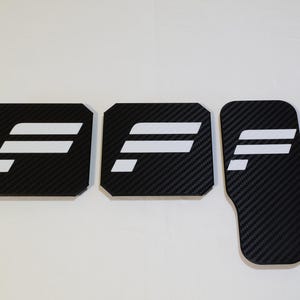 Op de afbeelding: Drie zwarte pedaalcovers met een koolstofvezeltextuur en een wit gestileerd letter F-logo. Twee van de covers zijn vierkant met afgeschuinde hoeken, terwijl de derde cover rechthoekig is met een afgeronde onderkant, alle met een consistent ontwerp.