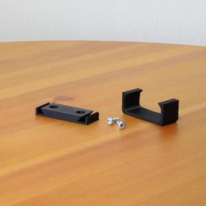 May include: Two black plastic mounting brackets with rectangular bases and U-shaped profiles, accompanied by two small silver metal screws. The components are arranged on a surface, showing the hardware needed for installation and secure attachment.