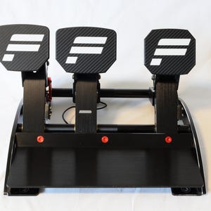 Op de afbeelding: Een zwarte racepedaalset met drie pedalen met een koolstofvezeltextuur en een wit F-logo op elk oppervlak. De set bevat een platte zwarte basisplaat, verticale metalen armen en drie kleine rode cirkelvormige accenten aan de onderkant.