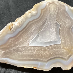 Puede incluir: Una rodaja de ágata pulida con bandas concéntricas de blanco translúcido, crema y gris claro. El centro contiene una pequeña cavidad de geoda cristalina con depósitos minerales brillantes que reflejan la luz y bordes naturales dentados.