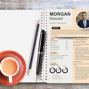 May include: A professional resume for a graphic designer, featuring a headshot, skills, and experience. A cup of coffee on a red saucer, a black pen, and a pencil rest on a wooden surface next to the open notebook.