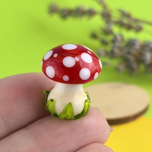 May include: A miniature glass mushroom with a red cap and white polka dots. The stem is off-white, and the base features green glass leaves. This decorative item is a whimsical addition to any collection.