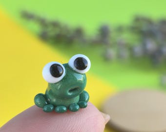 May include: A miniature green frog figurine with large, white-rimmed eyes and black pupils. The frog is perched on a fingertip, showcasing its small size. The background is a blend of yellow and green.