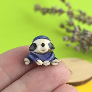 May include: A miniature figurine of a sloth wearing a blue hooded outfit. The sloth has a white face, black ears, and a small, round nose. The figurine is set against a green background with a sprig of lavender.