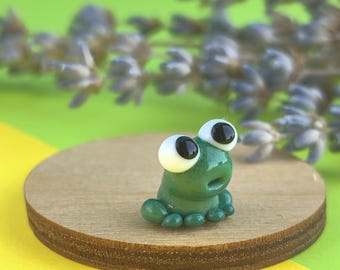 May include: A miniature green frog ornament with oversized eyes, set on a circular wooden base. The background features a blend of yellow and green hues, with a sprig of lavender. The frog is a charming decorative item.