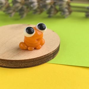 May include: A small, orange glass frog figurine with large, black-pupiled eyes. The frog is sitting on a round, wooden base. The background is a mix of green and yellow.