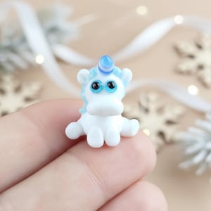 May include: A small, white unicorn figurine with a blue horn and mane. The unicorn has large, black-pupiled eyes and is posed with its arms outstretched. The background is a soft, blurred beige with decorative elements.