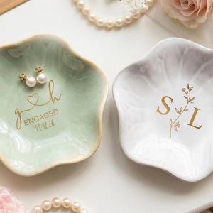Può includere: Due piattini portagioie in ceramica a forma di fiore con bordi smerlati. Il piattino a sinistra è verde salvia con testo dorato g h ENGAGED 11.12.26 e orecchini di perle. Il piattino a destra è bianco con testo dorato S L e un rametto floreale.