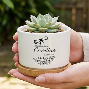May include: A white ceramic planter with a succulent plant, resting on a bamboo coaster. The planter is personalized with the name "Caroline", a graduation cap, and the text "Class of 2026". The succulent has pale green leaves.