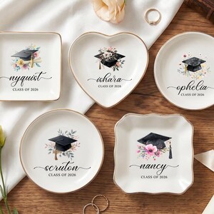 May include: A collection of personalized ceramic ring dishes in various shapes, each featuring a graduation cap design with floral accents. Names and "Class of 2026" are elegantly scripted on each dish, perfect for graduation gifts.