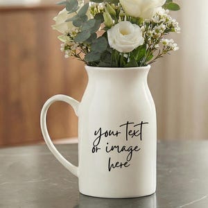 Puede incluir: Un jarrón blanco de cerámica estilo jarra con un asa curva, lleno de rosas blancas y vegetación. El jarrón tiene el texto "your Text or image here" en escritura negra. El jarrón está sobre una superficie gris.