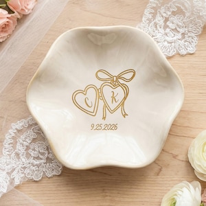 May include: A cream-colored, scalloped-edge ceramic trinket dish featuring a gold-colored graphic of two connected hearts with the letters C and K inside, tied with a bow, and the date 9.25.2026 printed in gold below the hearts.