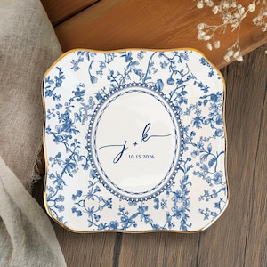 Puede incluir: Plato cuadrado de cerámica con diseño floral azul y borde dorado. El centro presenta un óvalo con las iniciales "j + k" y la fecha "10.15.2026" en escritura cursiva. El plato está sobre una superficie de madera.