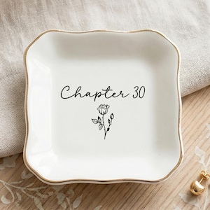 May include: A square white ceramic trinket dish with wavy edges and a gold-painted rim. The center features the black text "Chapter 30" written in a cursive script above a small, detailed black line drawing of a single rose with leaves.