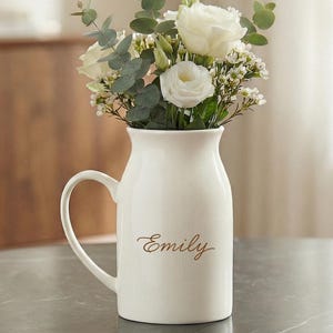 Puede incluir: Un jarrón de cerámica blanco con asa, lleno de rosas blancas y vegetación. El nombre "Emily" está escrito en cursiva en la parte delantera del jarrón. El jarrón está sobre una superficie oscura.