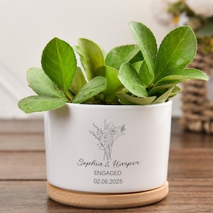 Könnte beinhalten: Ein weißer Keramik-Blumentopf mit floralem Design und den Namen "Sophia & Harper" mit dem Wort "ENGAGED" und dem Datum "02.06.2025". Der Topf enthält grüne Blätterpflanzen und steht auf einem runden Holzsockel.