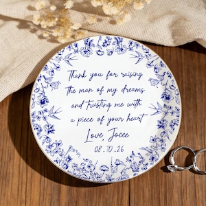 May include: A white ceramic plate with a blue floral design and gold trim. The plate has a handwritten message and the date 08.10.26. Two silver rings are next to the plate. The plate is on a wooden surface.