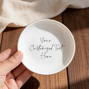 Op de afbeelding: Een kleine, ronde witte keramische schaal met een gouden rand, vastgehouden in een hand. De schaal heeft de tekst "Your Customized Text Here" in zwart schrift. De achtergrond omvat een houten oppervlak en een beige doek.