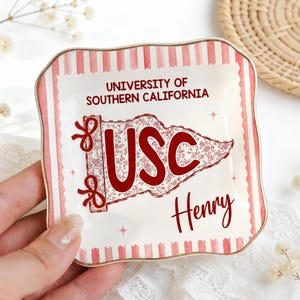 May include: A decorative ceramic trinket dish with a red and white striped border. The dish features the text "UNIVERSITY OF SOUTHERN CALIFORNIA" and a USC flag design in red. The name "Henry" is also written on the dish.