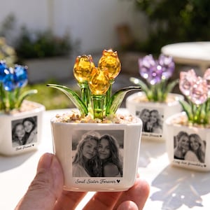 Pode incluir: Um vaso de cerâmica branco com uma foto de duas mulheres e o texto "Soul Sister Forever" contém tulipas de vidro amarelo com caules e folhas verdes. Outros vasos com flores azuis, roxas e rosas são visíveis.