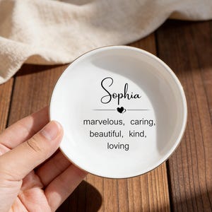May include: A white ceramic trinket dish with a gold rim. The dish features the name "Sophia" in elegant script, along with the words "marvelous, caring, beautiful, kind, loving". The dish is on a wooden surface.