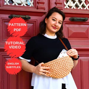 以下が含まれることがあります： 女性が、茶色の革のストラップが付いた、明るい茶色のテクスチャード加工されたかぎ針編みのクロスボディバッグを持っています。 バッグは三日月型で、ポケットに携帯電話が入っています。 画像には「PATTERN PDF」、「TUTORIAL VIDEO」、「ENGLISH SUBTITLES」というテキストが含まれています。
