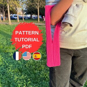 Könnte beinhalten: Ein leuchtend rosa gestrickter Schal hängt über dem Arm einer Person, die einen grauen Pullover und eine schwarze Jeans trägt. Das Bild enthält den Text "PATTERN TUTORIAL PDF" in einem roten Kreis sowie die Flaggen von Frankreich, den Vereinigten Staaten und Spanien.