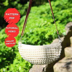 以下が含まれることがあります： クリーム色の、かぎ針編みのハンドバッグ。テクスチャード加工のボブルステッチのデザイン。茶色の革ストラップとシルバーカラーの金具が付いています。画像には「PATTERN PDF」、「TUTORIAL VIDEO」、「ENGLISH SUBTITLES」というテキストが含まれています。