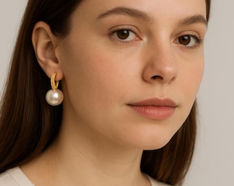 May include: A close-up of a person wearing a gold hoop earring with a large, white pearl pendant. The hoop is encrusted with small, sparkling details. The earring hangs from the ear, showcasing the pearl's smooth surface.