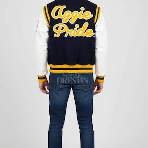 May include: Navy blue and white letterman jacket with "Aggie Pride" in yellow script on the back. The jacket has yellow and white striped accents and the word "Drestin" is printed on the bottom. The person is wearing blue jeans and white sneakers.