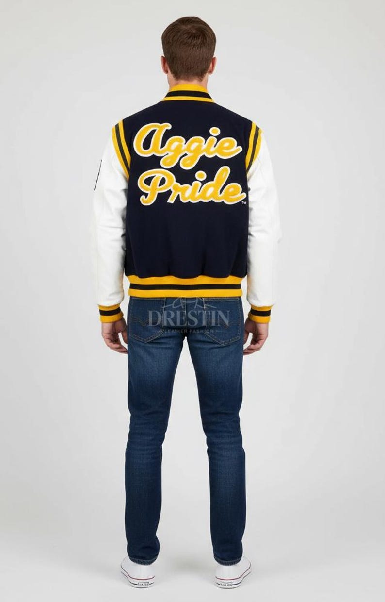 May include: Navy blue and white letterman jacket with "Aggie Pride" in yellow script on the back. The jacket has yellow and white striped accents and the word "Drestin" is printed on the bottom. The person is wearing blue jeans and white sneakers.