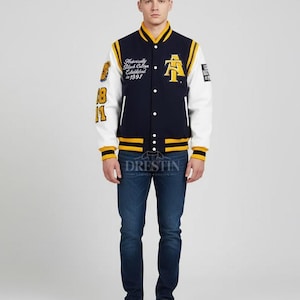 May include: Navy blue and white varsity jacket with yellow accents. Features the letter "A" and text on the chest and sleeve. The jacket has a button-up front and ribbed cuffs. The model is wearing blue jeans and white sneakers.